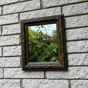 Puede incluir: Un espejo pequeño y rectangular con un marco oscuro y ornamentado. El espejo está montado en una pared de ladrillo blanco.