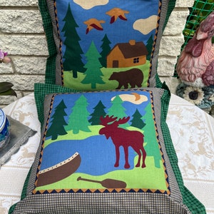 May include: Two decorative throw pillows with a woodland theme. The top pillow features a cabin, trees, a bear, and birds against a blue sky. The bottom pillow features a moose, a canoe, and a paddle against a green and blue background.