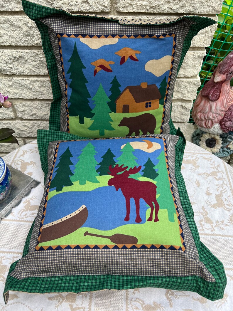 May include: Two decorative throw pillows with a woodland theme. The top pillow features a cabin, trees, a bear, and birds against a blue sky. The bottom pillow features a moose, a canoe, and a paddle against a green and blue background.