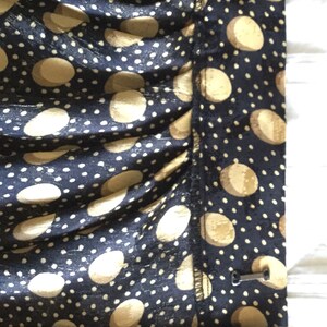 May include: Close-up of a navy blue fabric with a pattern of large, light tan circles and small white dots. The fabric is gathered, and a metal clasp is visible on the side. The overall look suggests a garment or textile.