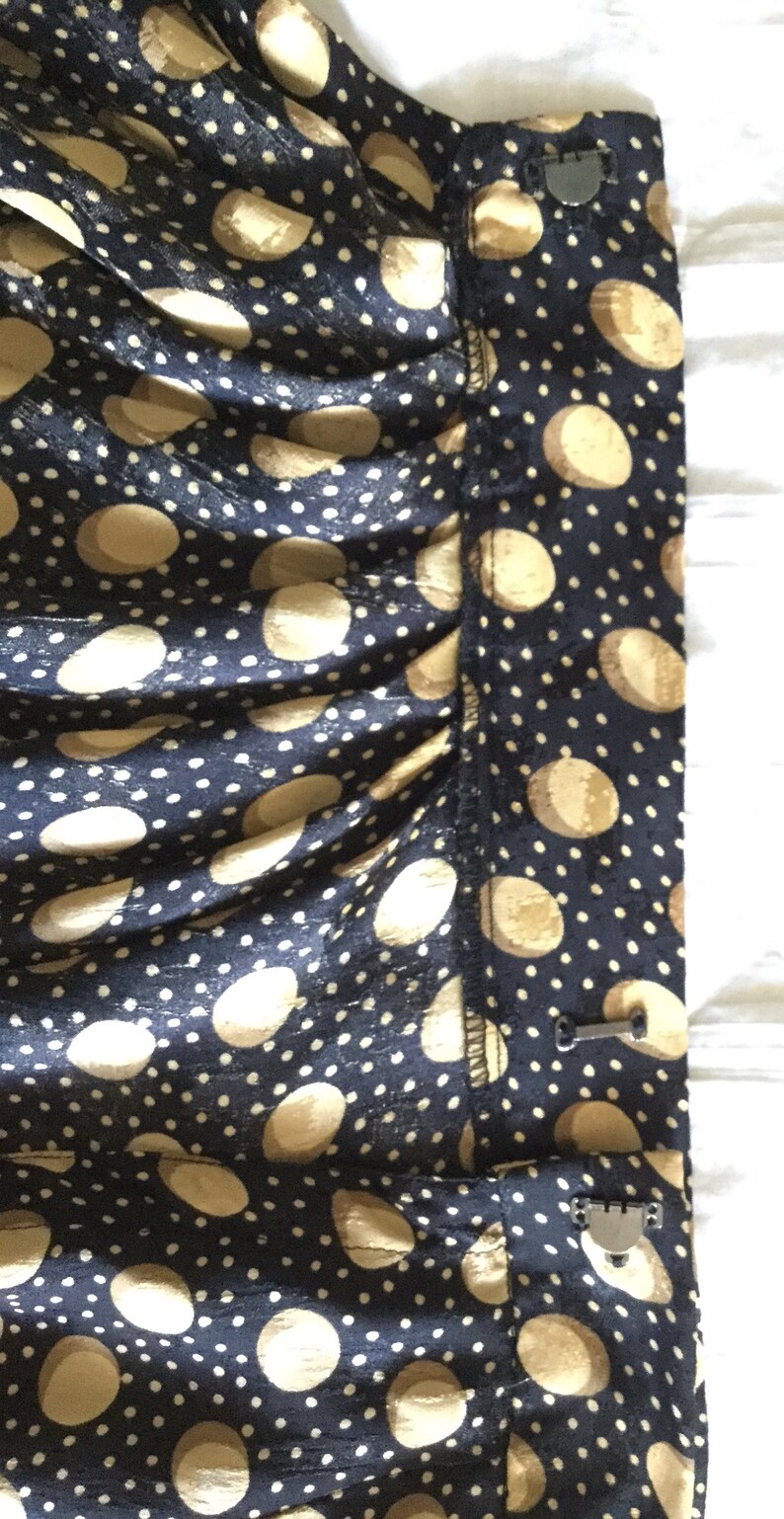 May include: Close-up of a navy blue fabric with a pattern of large, light tan circles and small white dots. The fabric is gathered, and a metal clasp is visible on the side. The overall look suggests a garment or textile.