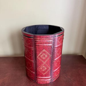 May include: A cylindrical red container with a black interior, designed to resemble a stack of books. The container features gold floral and diamond-shaped patterns, with black accents. The container is made of a sturdy material.