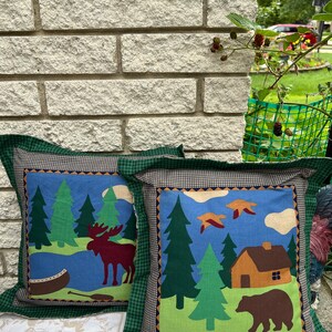 May include: Two decorative throw pillows with a woodland theme. One pillow features a moose and a canoe in a forest setting, while the other pillow features a bear and a cabin in a forest setting. Both pillows have a green, blue, and brown color scheme.