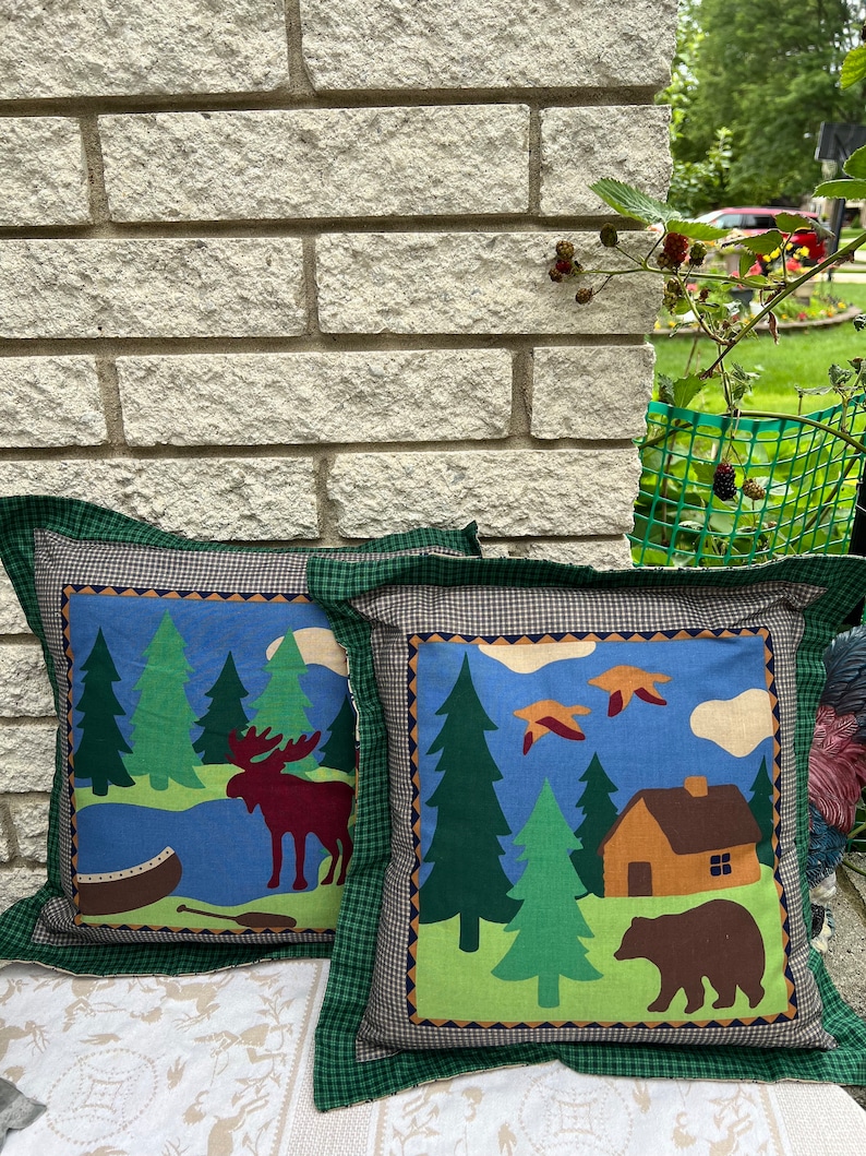 May include: Two decorative throw pillows with a woodland theme. One pillow features a moose and a canoe in a forest setting, while the other pillow features a bear and a cabin in a forest setting. Both pillows have a green, blue, and brown color scheme.