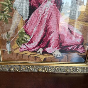 May include: A framed needlepoint tapestry depicting a figure in a flowing pink dress and white sleeves, seated on a stone ledge. The figure holds a small white object with a green stem. The artwork is encased in an ornate, metallic gold frame.