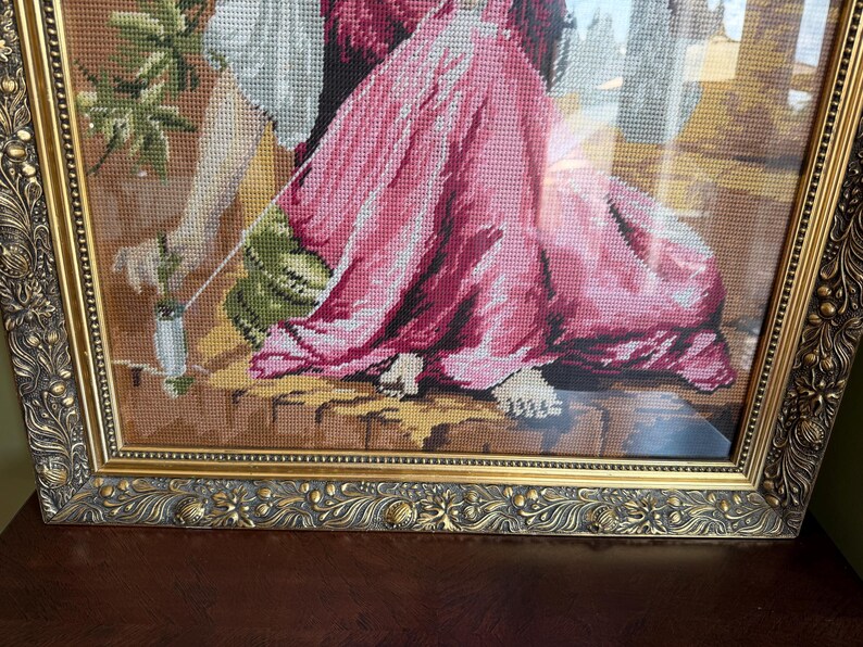 May include: A framed needlepoint tapestry depicting a figure in a flowing pink dress and white sleeves, seated on a stone ledge. The figure holds a small white object with a green stem. The artwork is encased in an ornate, metallic gold frame.