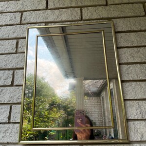 May include: A rectangular wall mirror featuring a double-layered gold-toned metal frame. The design consists of an outer rectangular border and an inner rectangular border, creating a clean, geometric look that reflects the surrounding outdoor area.