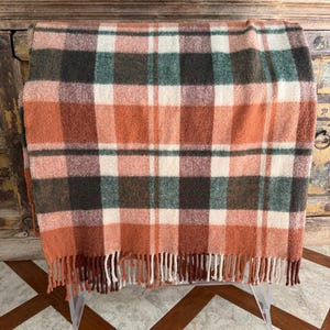 May include: A plaid blanket in shades of orange, green, and cream, with a fringed edge. The blanket is folded over a clear acrylic stand, showcasing its pattern. The blanket's design features a classic check pattern.