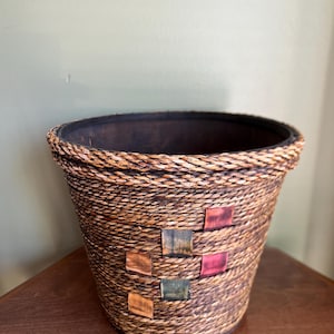 Może przedstawiać: Brązowy, pleciony koszyk na rośliny z kolorowymi akcentami z tkaniny. Koszyk jest okrągły i ma szeroki otwór.