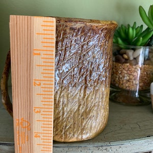 May include: A brown and white ceramic mug with a textured surface. The mug is approximately 5 inches tall.