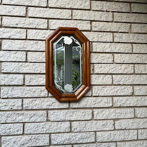 Może przedstawiać: Ośmiokątne lustro z brązową drewnianą ramą zamontowane na jasnoszarej ceglanej ścianie. Lustro ma wytrawione kwiatowe wzory i odbija otoczenie. Element dekoracyjny do wystroju wnętrz.