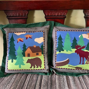 May include: Two decorative throw pillows with a woodland theme. One pillow features a brown bear and a cabin in a forest setting. The other pillow features a moose standing by a lake with a canoe.