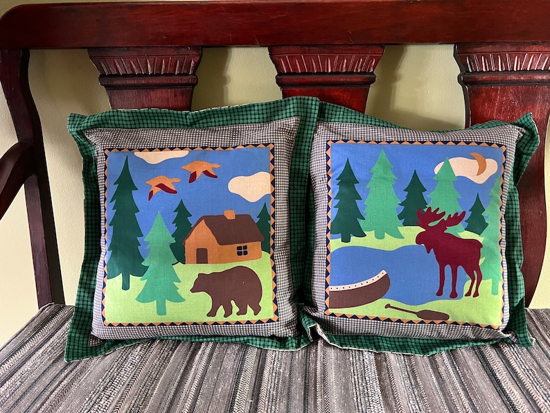 May include: Two decorative throw pillows with a woodland theme. One pillow features a brown bear and a cabin in a forest setting. The other pillow features a moose standing by a lake with a canoe.