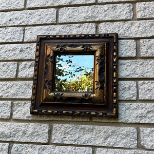 Può includere: Uno specchio piccolo e decorato con una cornice in legno marrone scuro e dettagli dorati. Lo specchio riflette una vista di alberi e un cielo blu.