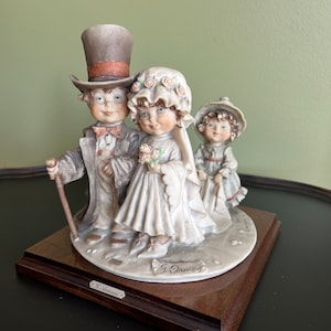 May include: A porcelain figurine set of three figures depicting a wedding ceremony. The bride and groom are dressed in traditional wedding attire, and the flower girl is wearing a white dress with a floral headband. The figurine is signed "G. Orsini" and "L. Armani" on the base.