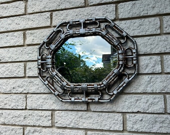 May include: An octagonal mirror with a silver metal frame. The frame has a chain-like design. The mirror is mounted on a light grey brick wall, reflecting the sky and trees.