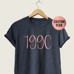May include: A dark heather grey t-shirt with a rose gold 1990 graphic. The shirt is hanging on a wooden hanger.