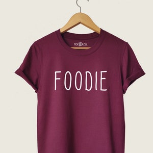 Puede incluir: Camiseta color burdeos con texto blanco que dice "FOODIE".
