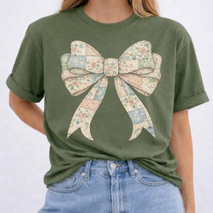 Puede incluir: Camiseta verde oliva con un gran diseño de lazo. El lazo está hecho de tela de retazos con patrones florales y geométricos en colores pastel. La camiseta tiene mangas cortas y un corte holgado.