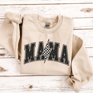 May include: A tan sweatshirt with a black and white checkered lightning bolt graphic and the word "MAMA" in a vintage style font.