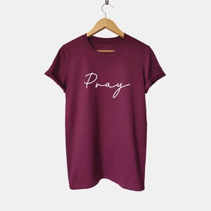 May include: Burgundy t-shirt with the word "Pray" written in white cursive script.