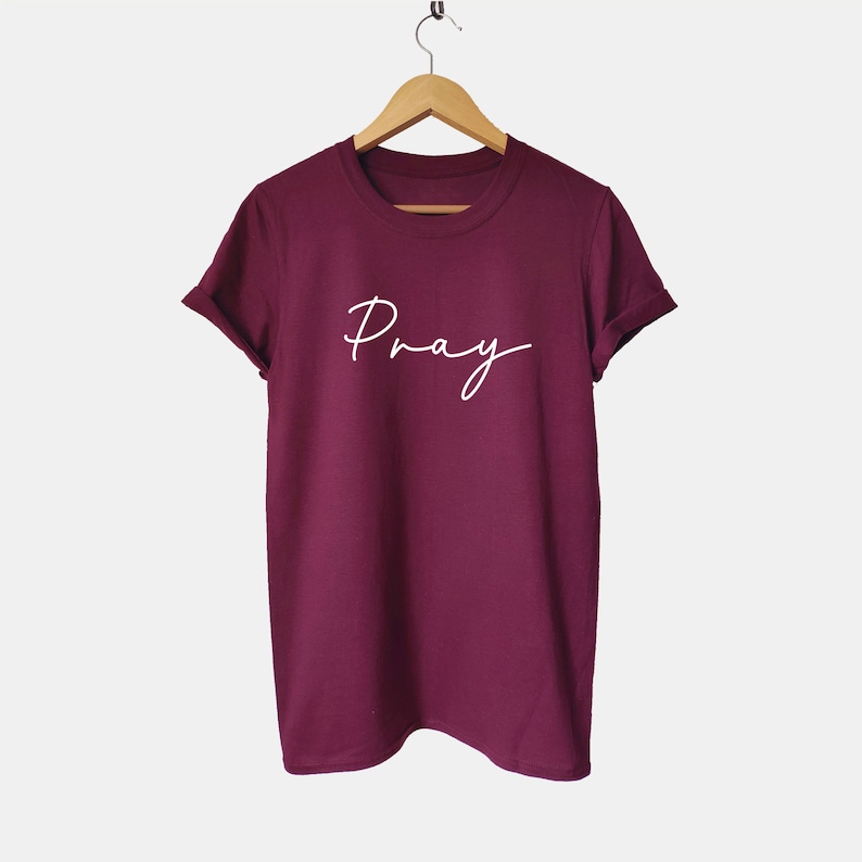 May include: Burgundy t-shirt with the word "Pray" written in white cursive script.