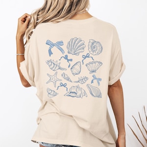 Op de afbeelding: Een beige T-shirt met een blauw schelp- en zeesterontwerp. Het ontwerp bevat een verscheidenheid aan schelpen, zeesterren en strikken.