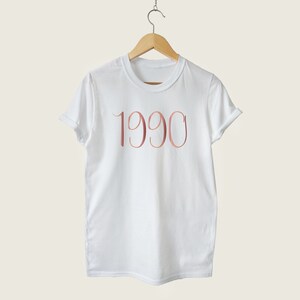 May include: White t-shirt with the year 1990 printed in rose gold on the front.