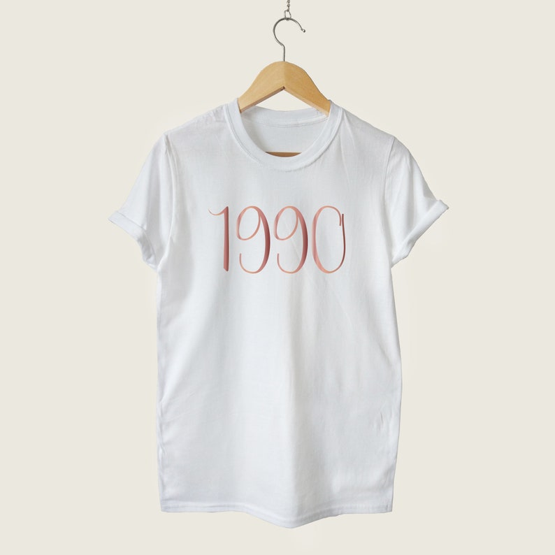 May include: White t-shirt with the year 1990 printed in rose gold on the front.