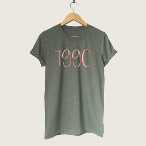 May include: A dark green t-shirt with a rose gold 1990 printed on the front.