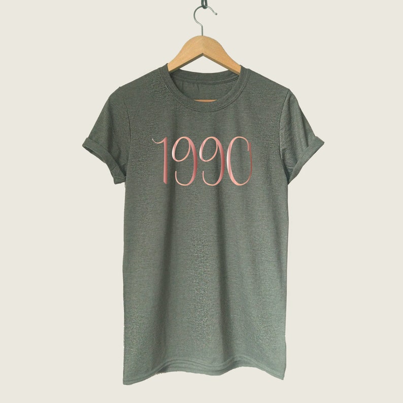 May include: A dark green t-shirt with a rose gold 1990 printed on the front.