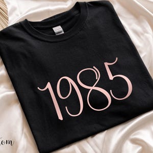 May include: Black t-shirt with a pink 1985 graphic printed on the front.