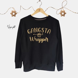 Puede incluir: Sudadera negra con el texto "Gangsta Wrapper" en letras doradas. El texto está estilizado con un pequeño gráfico de caja de regalo.