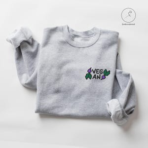 Op de afbeelding: Een grijs sweatshirt met het woord "VEGAN" geborduurd op de voorkant, omringd door kleine geborduurde paarse en groene groenten.