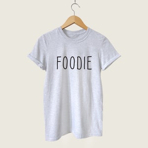 Puede incluir: Una camiseta gris con la palabra "FOODIE" impresa en negro en la parte delantera.