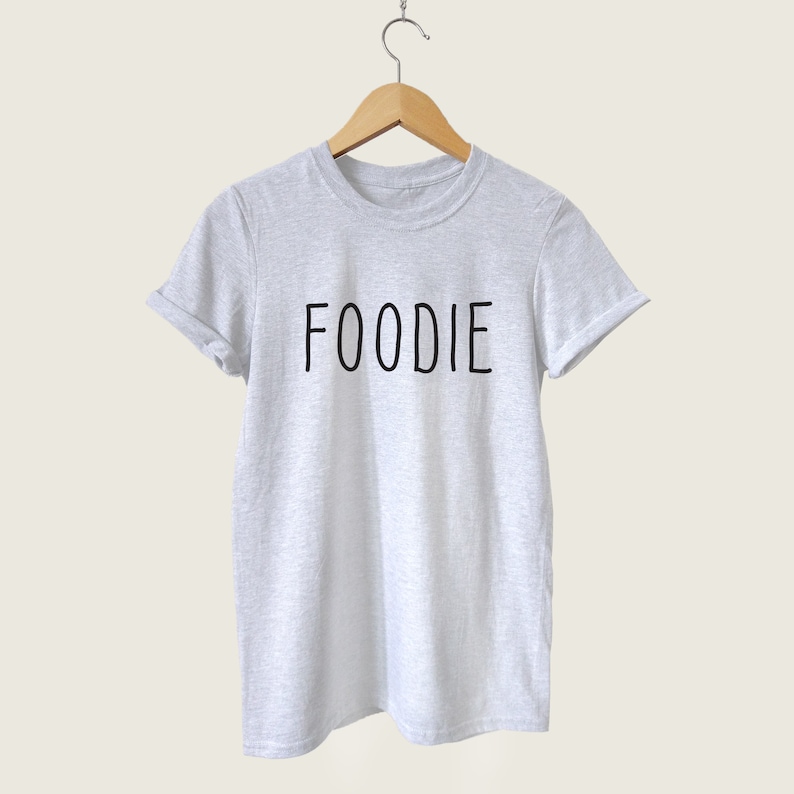 Puede incluir: Una camiseta gris con la palabra "FOODIE" impresa en negro en la parte delantera.