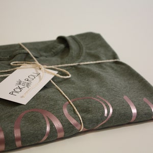 May include: A folded green t-shirt with a rose gold "You" design. The shirt is tied with twine and has a white tag that reads "Pick and Roll".
