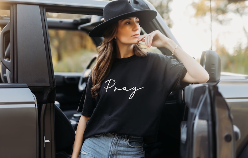 May include: A woman wearing a black t-shirt with the word "Pray" written in white script. She is standing next to a gray car with the door open.