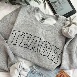 Könnte beinhalten: Ein grauer Pullover mit dem Wort "TEACH" in schwarzen umrandeten Buchstaben.
