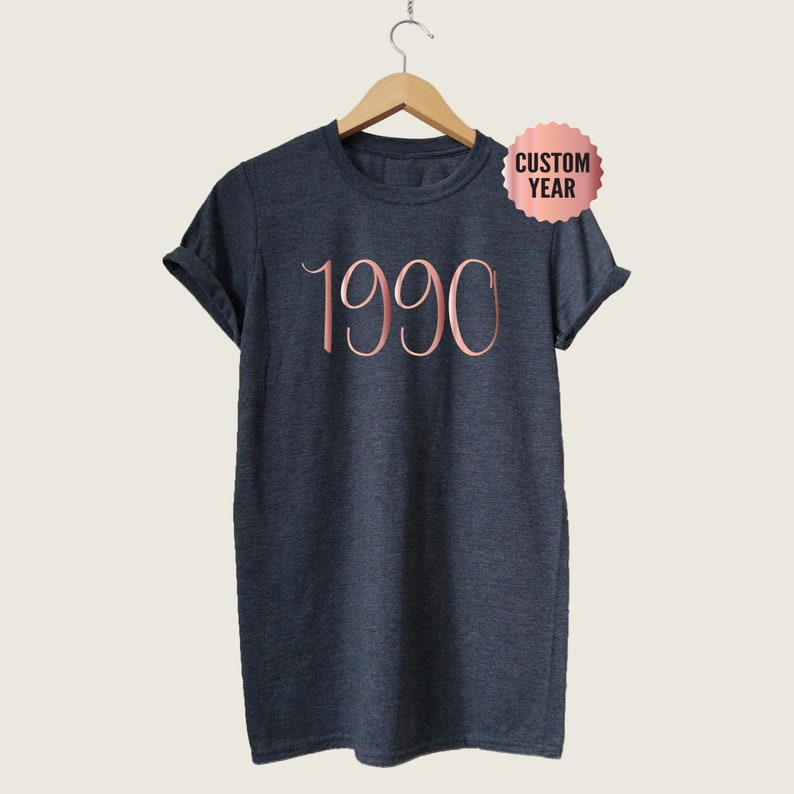 May include: A dark heather grey t-shirt with a rose gold 1990 graphic. The shirt is hanging on a wooden hanger.
