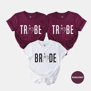 Op de afbeelding: Drie T-shirts met een bruid en bruidsmeisjes thema. Het bruidsshirt is wit en zegt "BRIDE" met een middelvinger en ring erop. De twee bruidsmeisjesshirts zijn bordeauxrood en zeggen "TRIBE" met een middelvinger en ring erop.