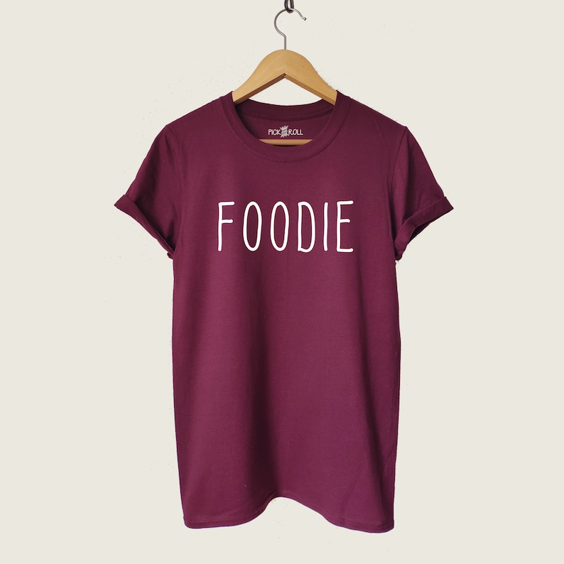 Puede incluir: Camiseta color burdeos con texto blanco que dice "FOODIE".