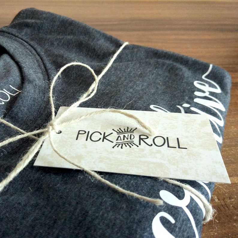 Puede incluir: Una camiseta gris con texto blanco que dice "Pick and Roll" en una etiqueta marr&oacute;n. La etiqueta est&aacute; atada a la camiseta con un cordel.