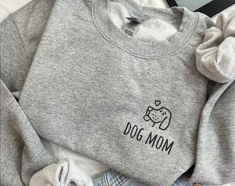 May include: A heather gray crewneck sweatshirt featuring a black line art graphic of a dog face with a small heart above it and the text DOG MOM printed in a simple sans-serif font on the left chest area of the garment.