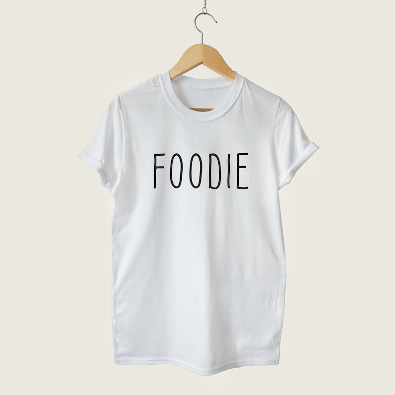 Puede incluir: Camiseta blanca con la palabra "FOODIE" impresa en negro en la parte delantera.
