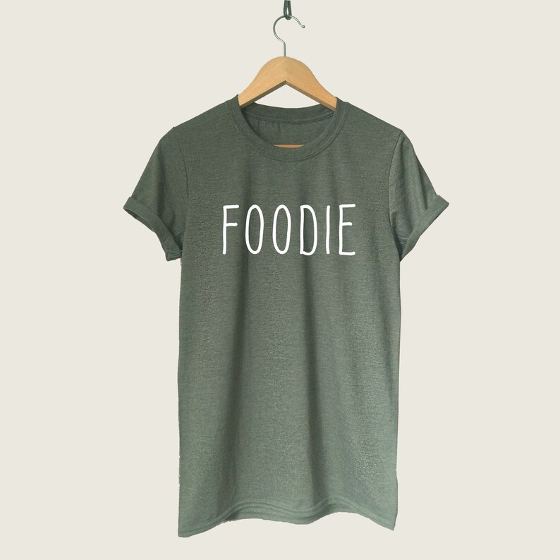 Puede incluir: Una camiseta verde con la palabra "FOODIE" impresa en blanco en la parte delantera.