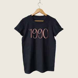 May include: Black t-shirt with a rose gold 1990 graphic.