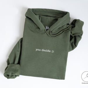May include: An olive green hoodie with a drawstring hood and long sleeves. The phrase "you decide :)" is embroidered in white on the front. The hoodie is made of a soft material and is suitable for casual wear.