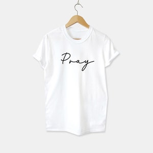 May include: White t-shirt with the word "Pray" written in black cursive script.