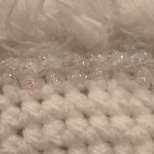 May include: Close-up of a textured, off-white crochet fabric with a layer of iridescent beads and a fluffy, white faux fur trim. The crochet has a distinct stitch pattern.